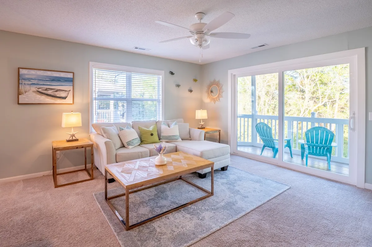 A comfortable sectional sofa anchors the living room, with a coffee table set for casual gatherings. The space is brightened by natural light streaming through large windows, and a glimpse of bright blue chairs on the adjacent porch hints at seamless indoor-outdoor living. The inviting ambiance makes it perfect for relaxing with a book, enjoying morning coffee, or hosting friends.
