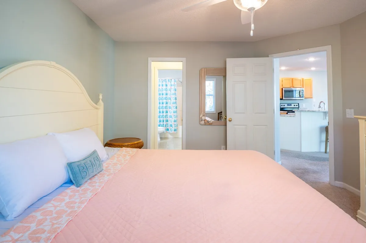 A cozy bedroom view featuring a bed with a soft pink quilt, a mirror, and doorways leading to the bathroom and kitchen. The soft lighting and neutral colors create an inviting and restful atmosphere.