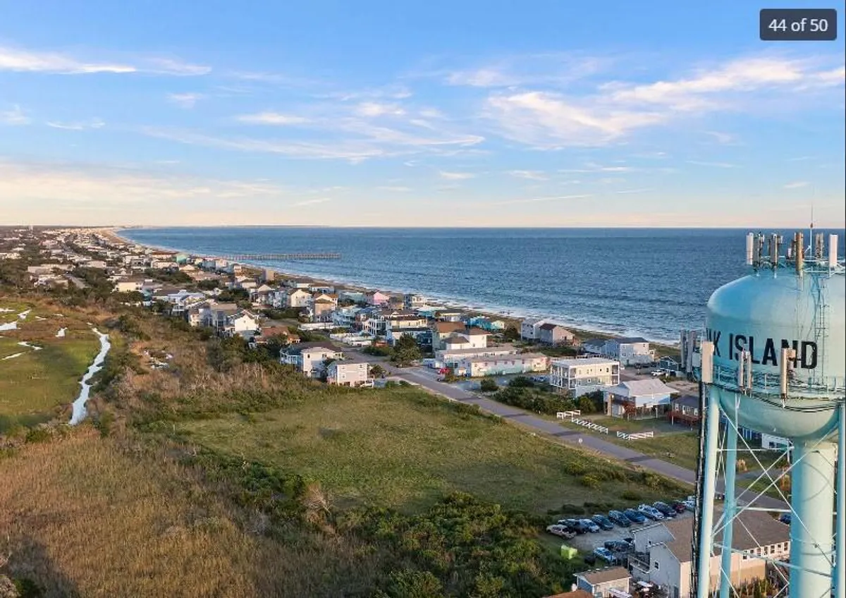 A wide view overlooking the beach, ocean, water tower, and surrounding houses. The sandy shoreline stretches out, inviting beach walks, sunbathing, and family picnics. Coastal roads and nearby homes evoke a sense of easy access to both private and public beach adventures.