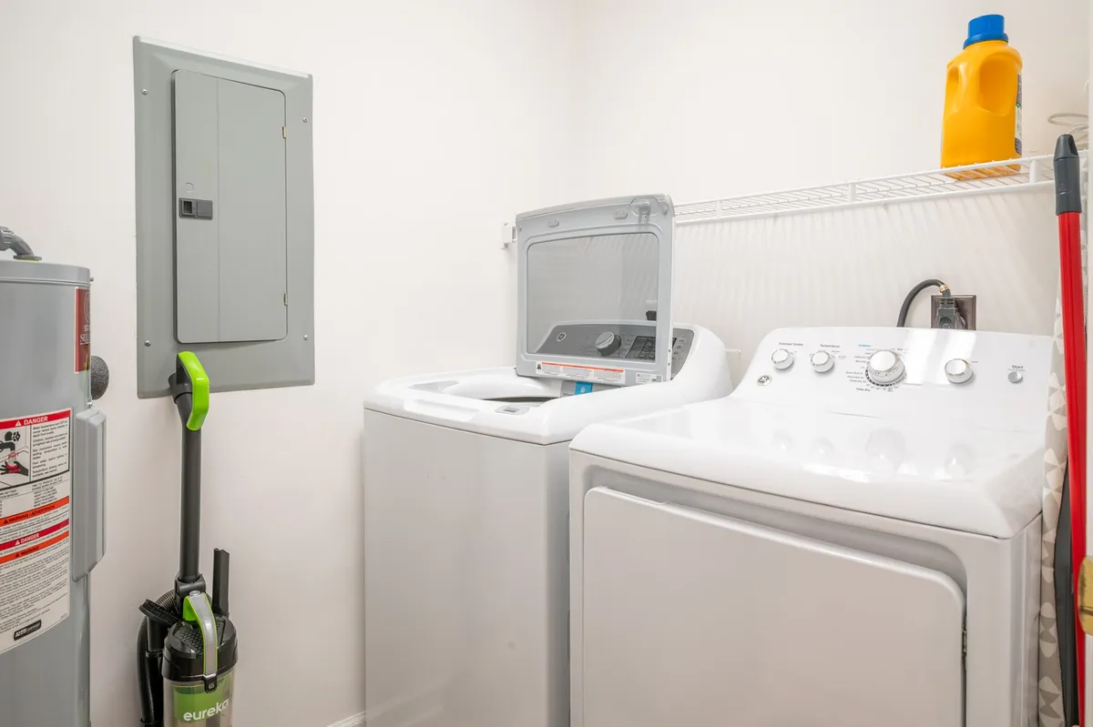 A compact but well-organized laundry area featuring a washing machine, dryer, detergent bottle, utility panel, and a vacuum cleaner. The space is practical and accessible, making laundry chores easy and efficient for guests.