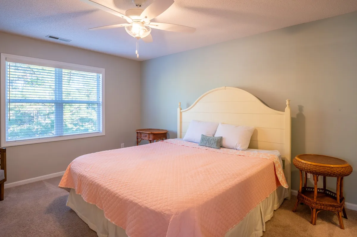 A spacious, serene bedroom featuring a large bed dressed in a pink quilt, flanked by two bedside tables. Natural light streams through a window with blinds, creating an airy and restful environment.