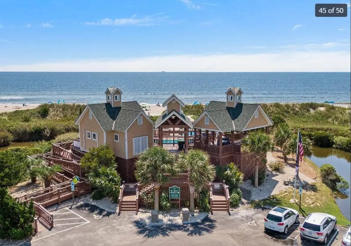The image features a cheerful scene with a beach house structure set against the backdrop of the ocean and sandy beach. Palm trees frame the area, while a parking lot ensures easy access for visitors. The lively atmosphere suggests family outings, swimming, sunbathing, and beach games, with community events often hosted at this vibrant location.