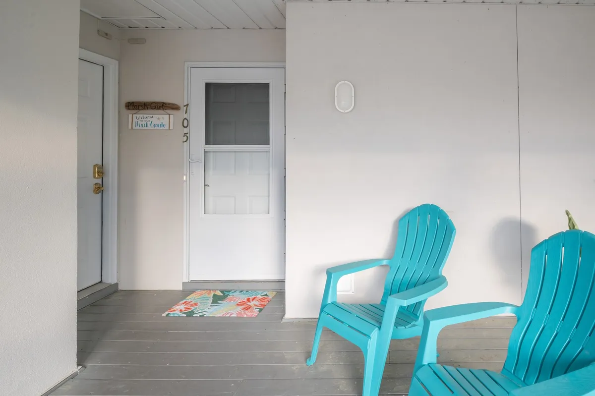 The image shows a welcoming exterior with turquoise chairs placed next to a door featuring a cheerful welcome mat and a door sign. The setup is inviting, suggesting a relaxed outdoor space perfect for unwinding and socializing. The chairs are positioned to enjoy the view and the ambiance of the condo’s entryway.