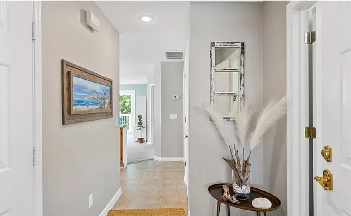 A calm and inviting entryway with a framed beach scene painting, a decorative mirror, a vase with pampas grass, and a small side table. The design sets a welcoming tone as you enter the home, reflecting thoughtful details for guests.