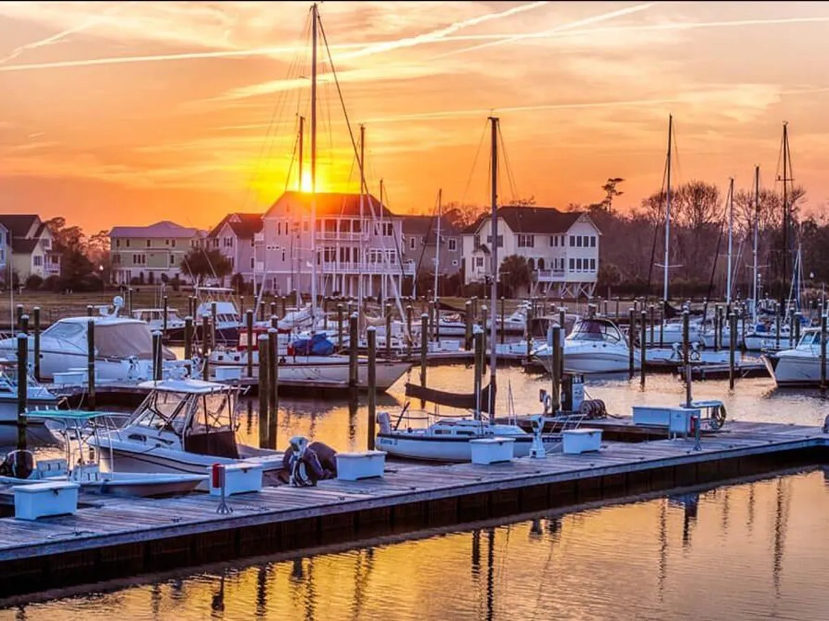 A serene waterfront scene with sailboats docked at sunset, water reflecting vibrant colors, and houses lining the shore. The atmosphere is peaceful, with opportunities for boating, fishing, or simply enjoying the view as the sun sets.