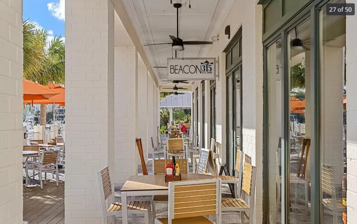 An inviting restaurant with prominent signage, shaded outdoor seating, and decorative potted plants, suggesting a lively yet relaxed dining atmosphere. The scene hints at social gatherings, people-watching, and the enjoyment of local cuisine in a scenic setting.