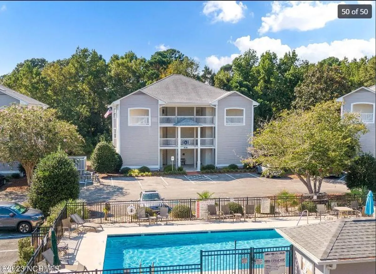 The image showcases the condo's inviting exterior set against lush green landscaping, with a sparkling swimming pool as the centerpiece. Comfortable outdoor seating is arranged around the pool, creating a relaxed atmosphere perfect for sunbathing, swimming, or gathering with friends and family. The scene exudes a welcoming, sociable vibe ideal for vacationers seeking both relaxation and connection.