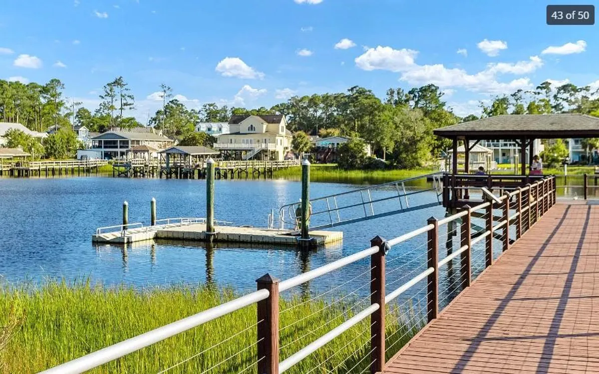 A picturesque dock extending over calm water, surrounded by greenery and nearby houses. The view suggests a relaxed environment ideal for boating, fishing, picnicking, or simply strolling along the waterfront.